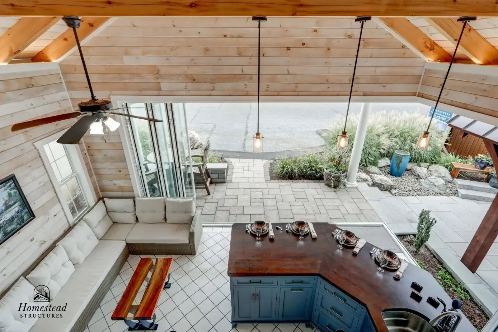 Interior view of a kitchen with large windows, gray cabinets with gold handles, a wooden countertop, and a small sink. The ceiling is wood-paneled with a hanging light fixture.
Outside the windows, there is a patio with a wooden pergola and neighbori