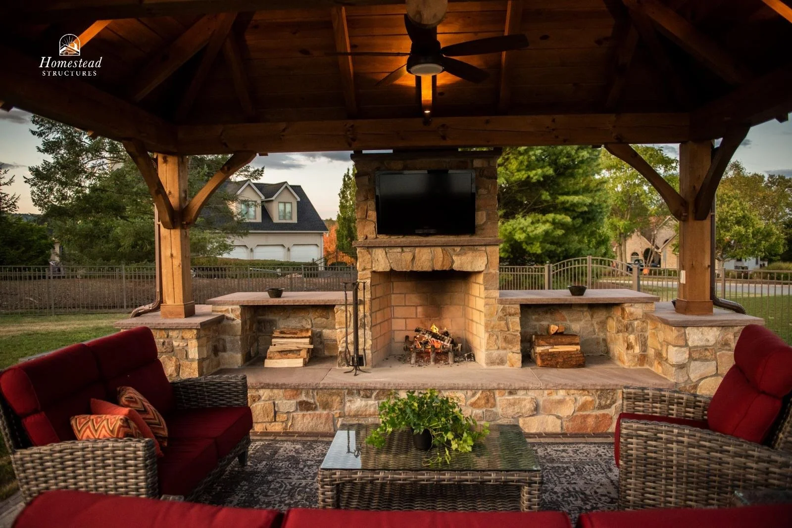 Outdoor covered patio with stone fireplace, TV mounted above, wicker furniture with red cushions, a glass-top coffee table with a plant, and a view of a suburban neighborhood with trees and houses.