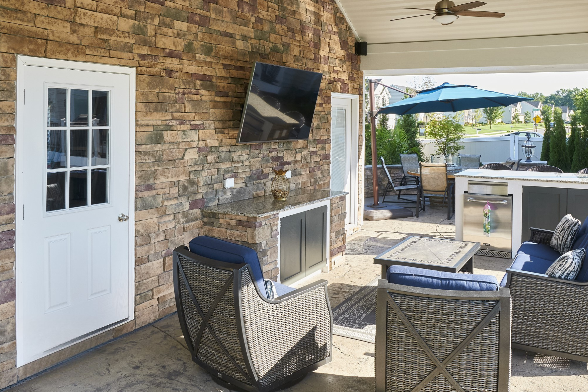 Outdoor patio with wicker furniture, blue cushions, a wall-mounted TV, and a dining area with a large blue umbrella. There's a brick wall and a small bar area with a mini fridge.