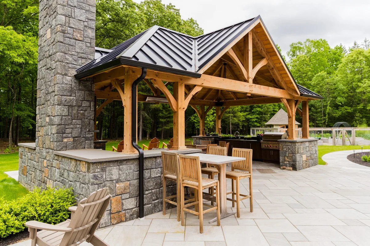 Outdoor kitchen with stone and wood structure, bar seating, and a barbecue grill, surrounded by greenery and trees.