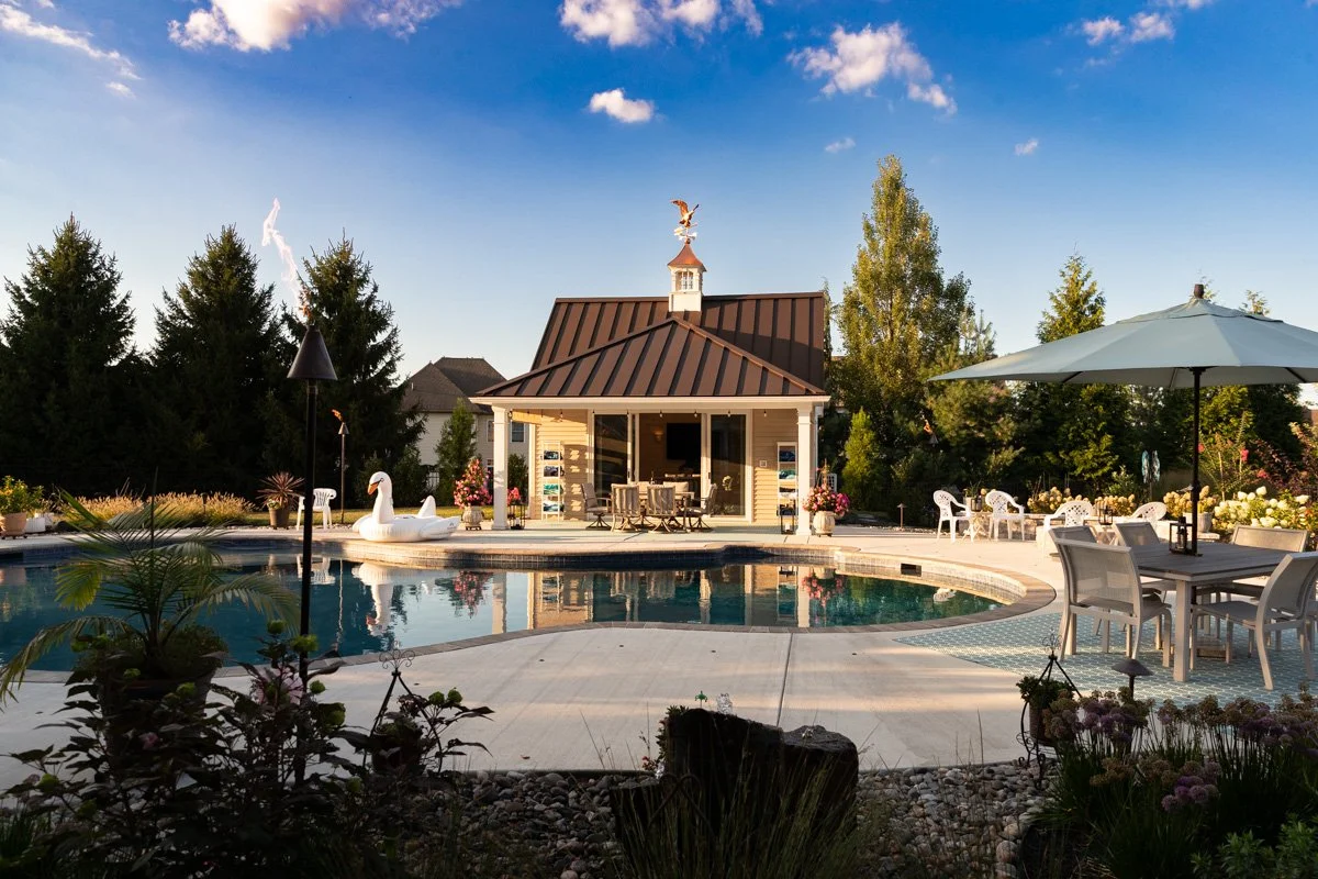 A backyard with a swimming pool, surrounded by outdoor furniture, plants, and trees. There is a small house with a porch and a weather vane on the roof, and a floating swan pool float in the water. The scene is sunny with a blue sky and a few clouds.