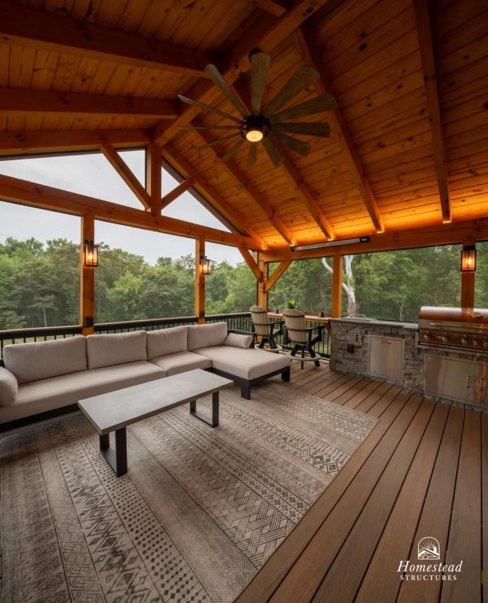 Covered outdoor porch with wood siding, ceiling, and flooring. Features a ceiling fan, a large beige sectional sofa, a rectangular coffee table, and a built-in outdoor kitchen with stone accents and a grill. Large windows overlook greenery.