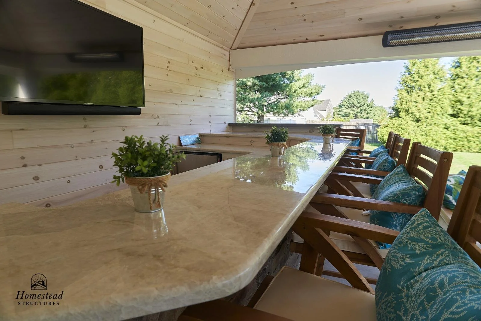 Indoor outdoor kitchen with a marble countertop bar, wooden chairs with blue cushions, a wall-mounted TV, and a large window overlooking trees and a yard.