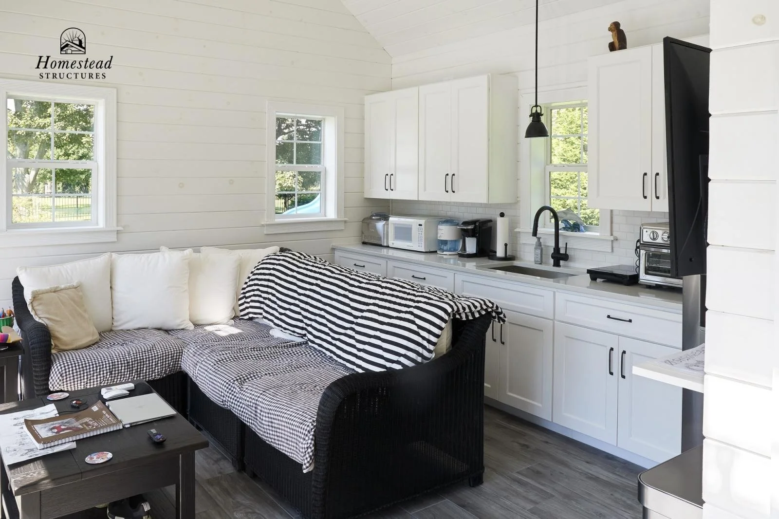 Living room kitchen with white cabinets, black countertop appliances, black wicker sectional with black and white striped throw blanket, three windows with green trees outside, and a black hanging pendant light.