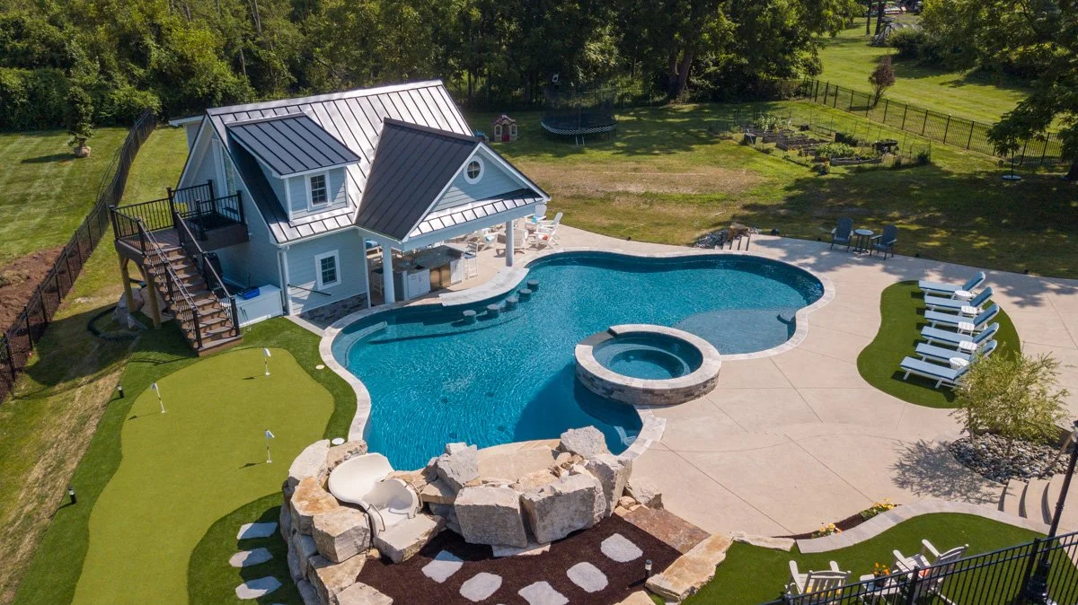 A backyard with a swimming pool, hot tub, and a pool house. The pool area has lounge chairs, a putting green, and surrounded by a black fence with trees and garden in the background.