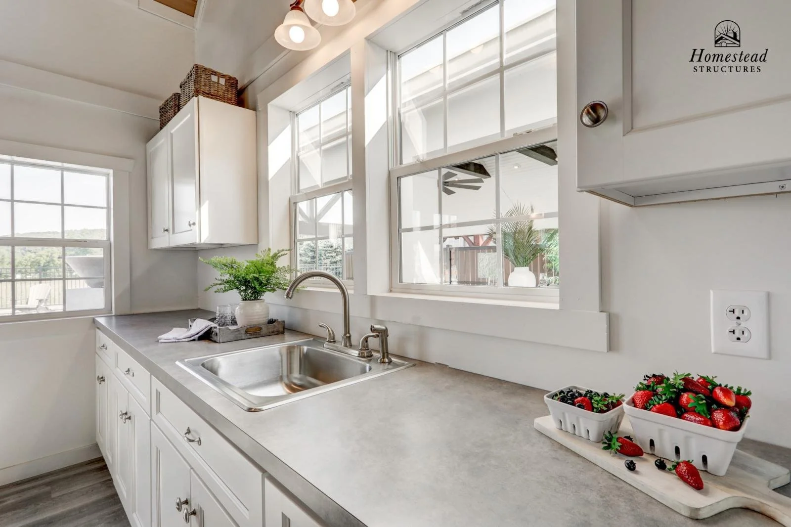 A bright kitchen with white cabinets, a sink, and large windows. Bowls of strawberries and mixed berries are on the countertop.