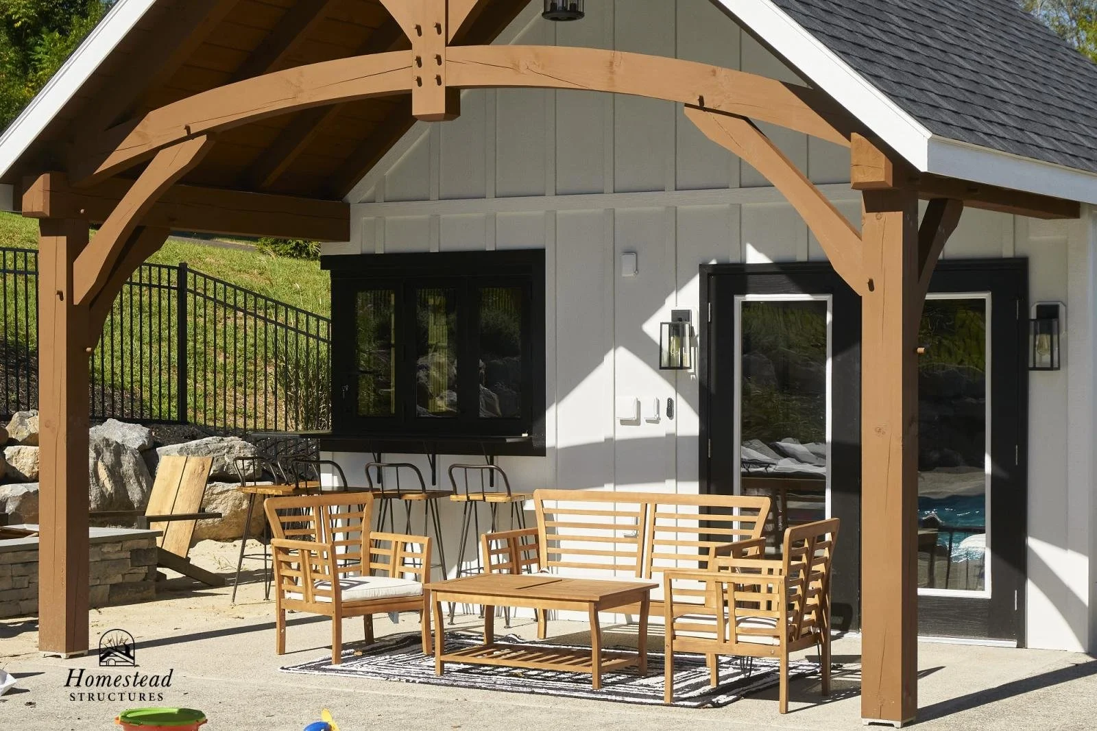 Outdoor patio with wooden furniture under a wooden pergola in front of a house. The house has white siding, black window and door frames, and exterior light fixtures. There is a grassy hillside with rocks and a fence in the background.