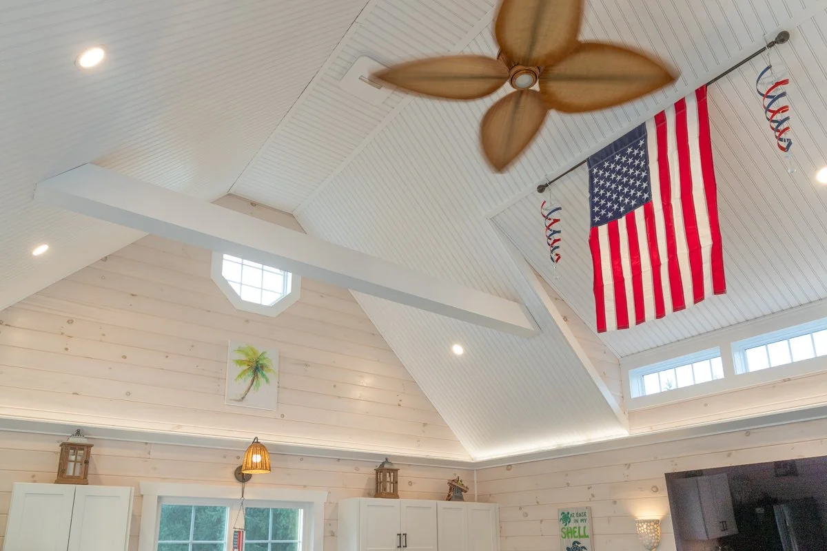 Interior of a bright room with a vaulted ceiling, an American flag hanging from the ceiling, a ceiling fan with wooden blades, and tropical-themed decorations on the walls.