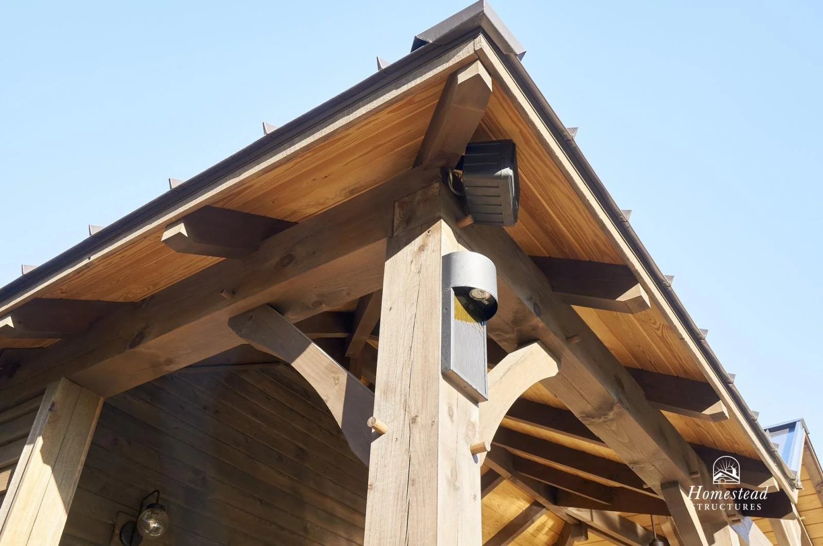 Close-up of the corner of a wooden porch roof with lighting fixtures and a security camera, showing weathered wood and clear blue sky.