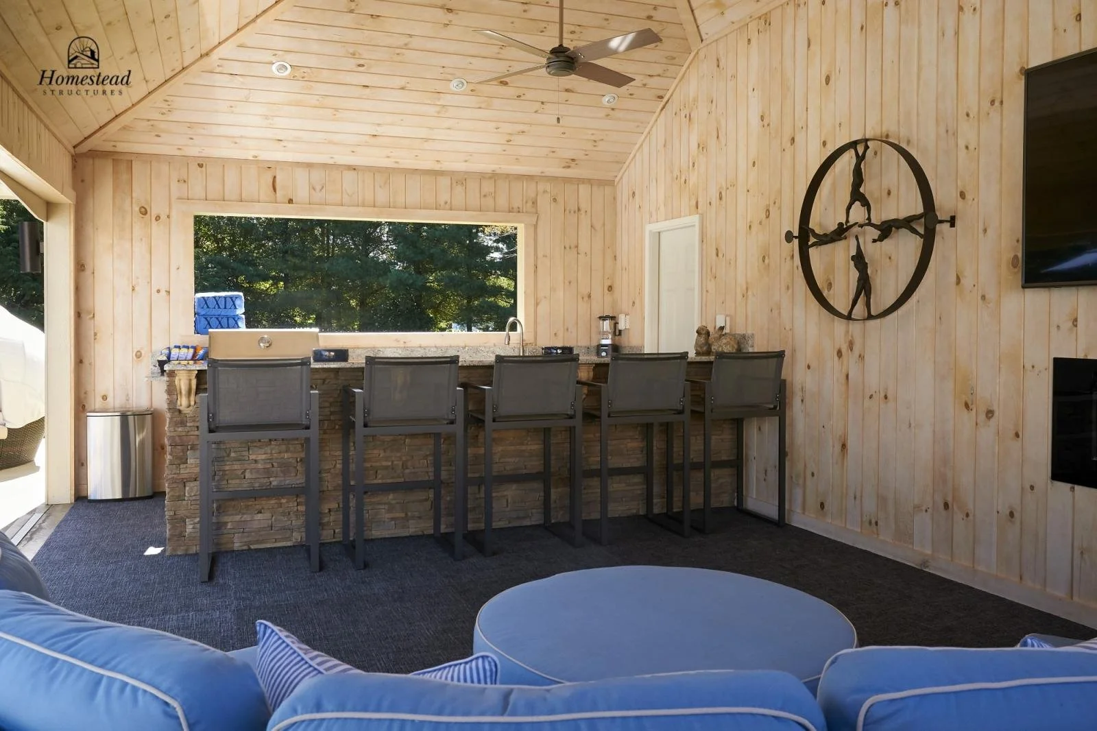 Interior of a wooden outdoor bar area with four bar stools, a stone bar counter, a large window, a ceiling fan, wall clock, and a flat-screen TV on the wall.