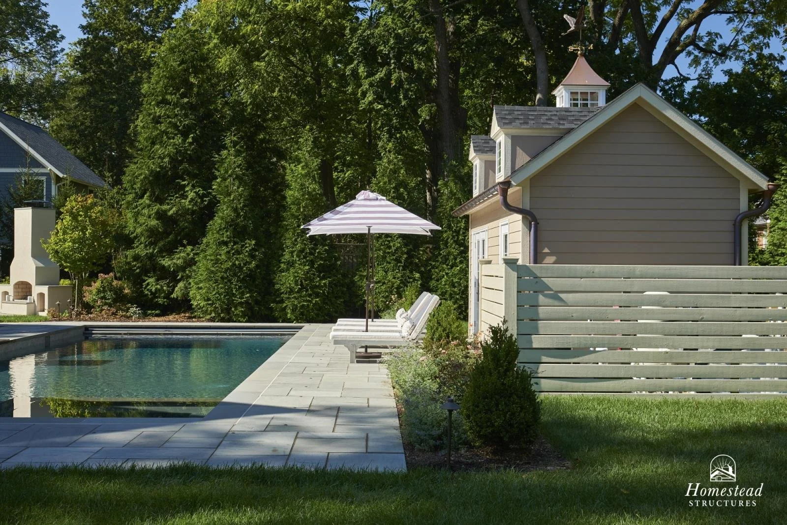 Backyard with a swimming pool, lounge chairs with a striped umbrella, small garden, wooden fence, and a small beige house with a cupola, surrounded by tall trees.