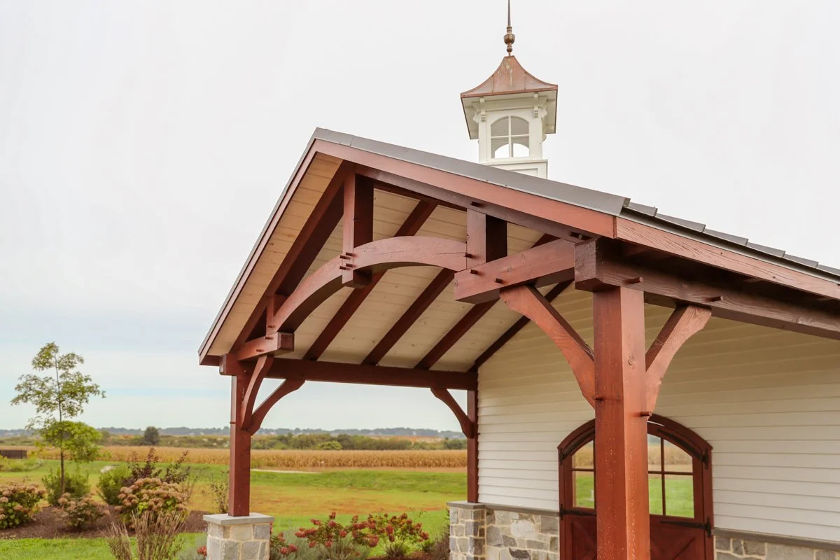 A rural building with a steep roof, wooden beams, a small tower on top, and green landscape in the background.