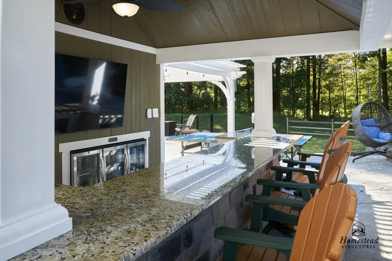 Outdoor patio with bar seating, flat-screen TV, and porch swing overlooking a wooded yard.
