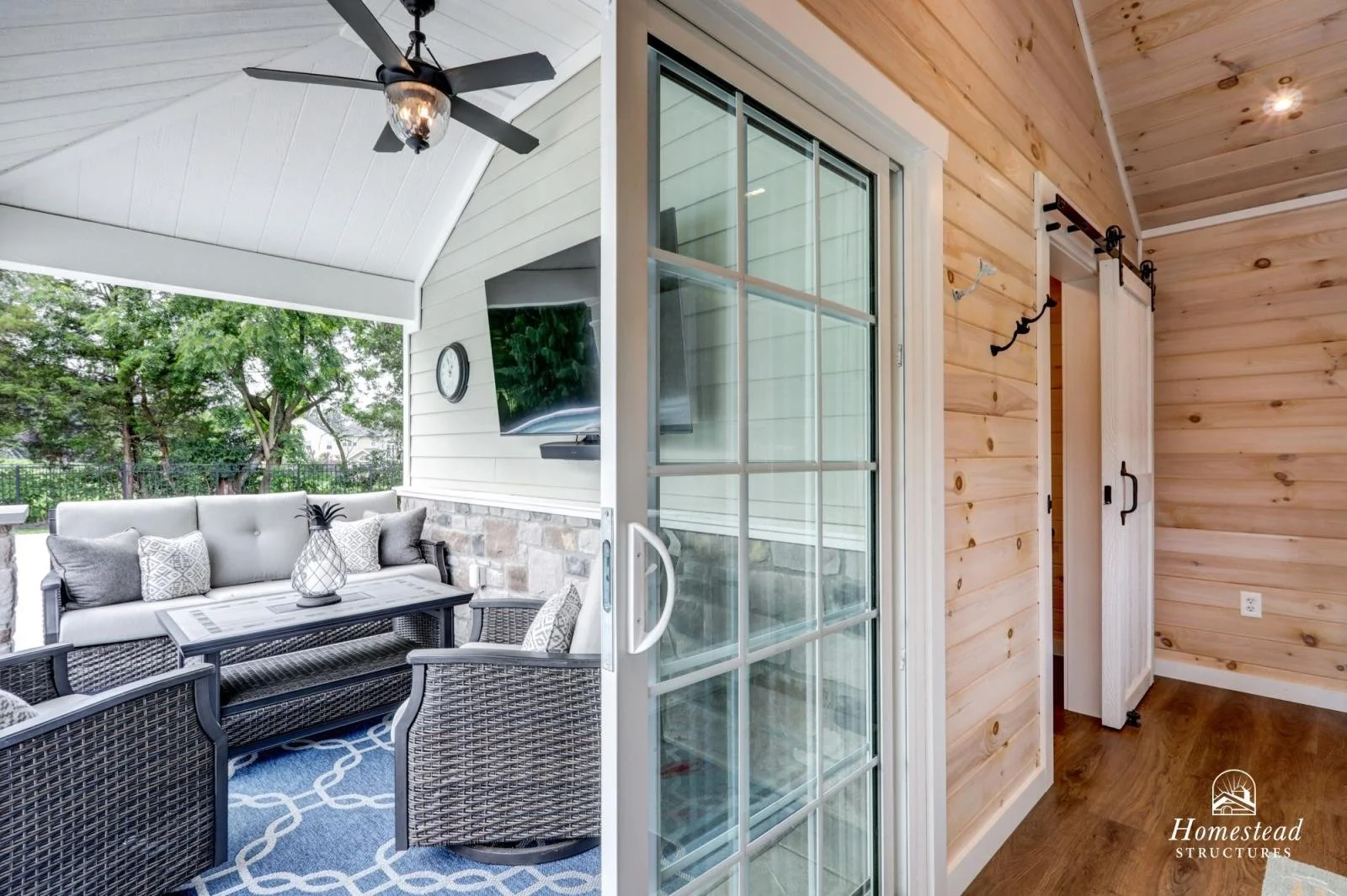 Interior view of a porch with wicker furniture and outdoor scenery, viewed from inside a room with wood paneling and sliding glass doors.