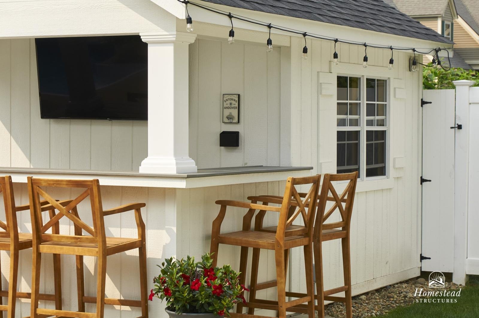 Outdoor patio area of a backyard with a bar counter, three wooden chairs, a potted plant with red flowers, a wall-mounted TV, and string lights overhead. The house exterior is painted white with a window and a door.