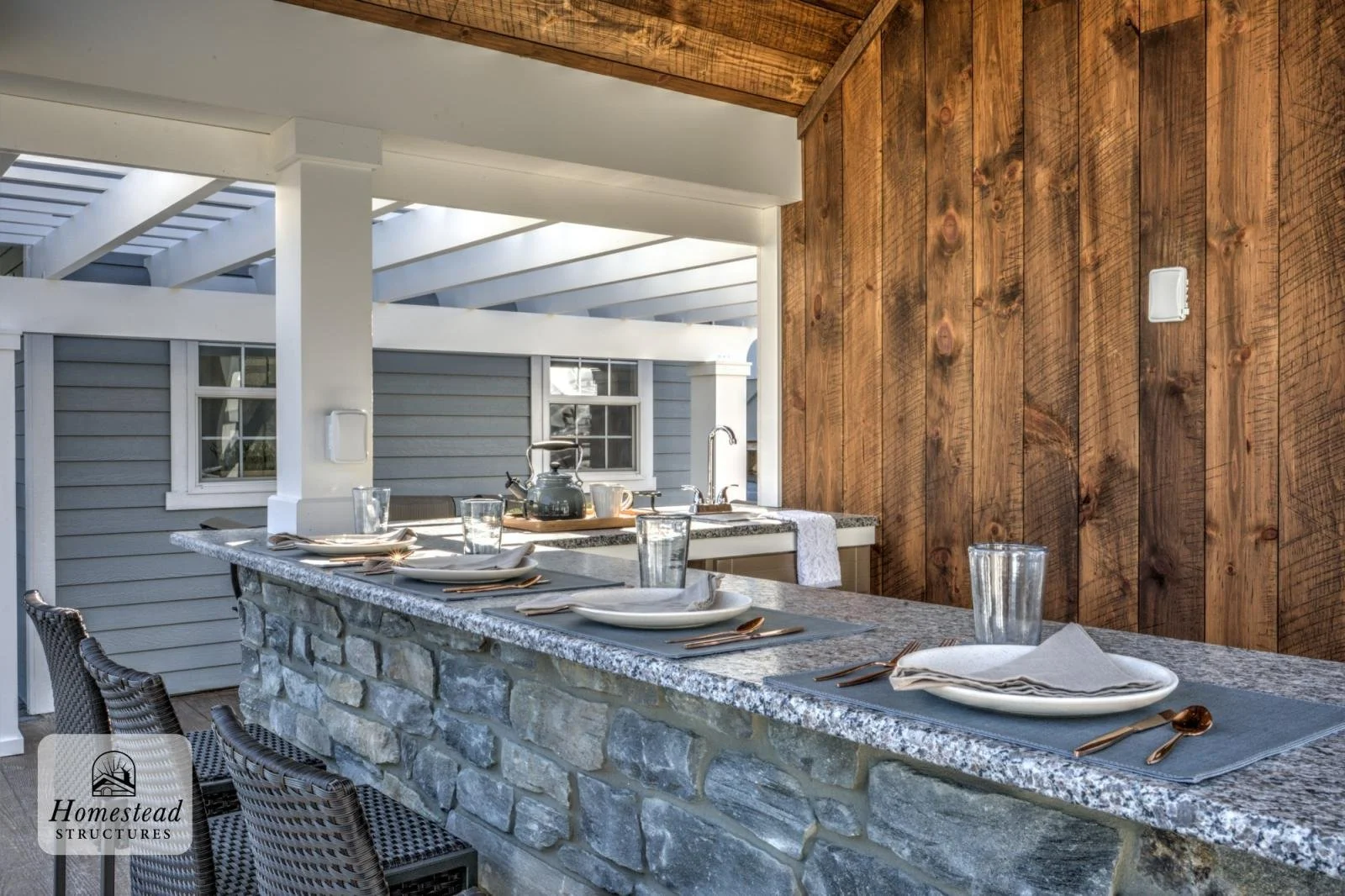 Outdoor dining area with a stone counter, set with plates, glasses, and utensils, featuring a wooden wall backdrop.