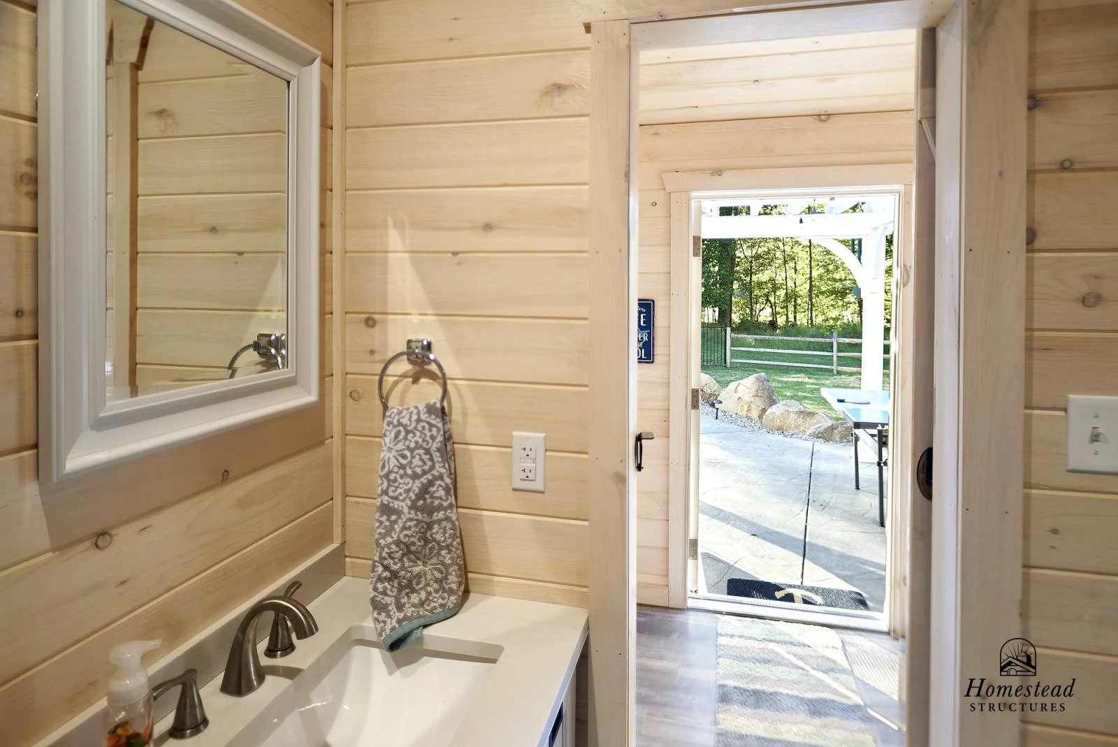 Interior of a small wooden bathroom with a sink, mirror, and towel rack, leading to an outdoor space with rocks, trees, and a patio.