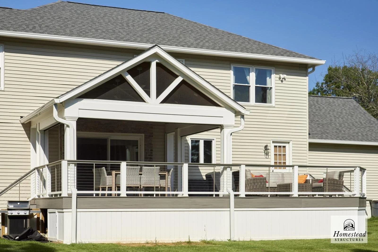 Backyard with a large wooden deck connected to a beige house with vinyl siding, multiple windows, and a gable roof. The deck has outdoor furniture, including chairs, a table, and a seating area with cushions. A grill is visible on the left side of th