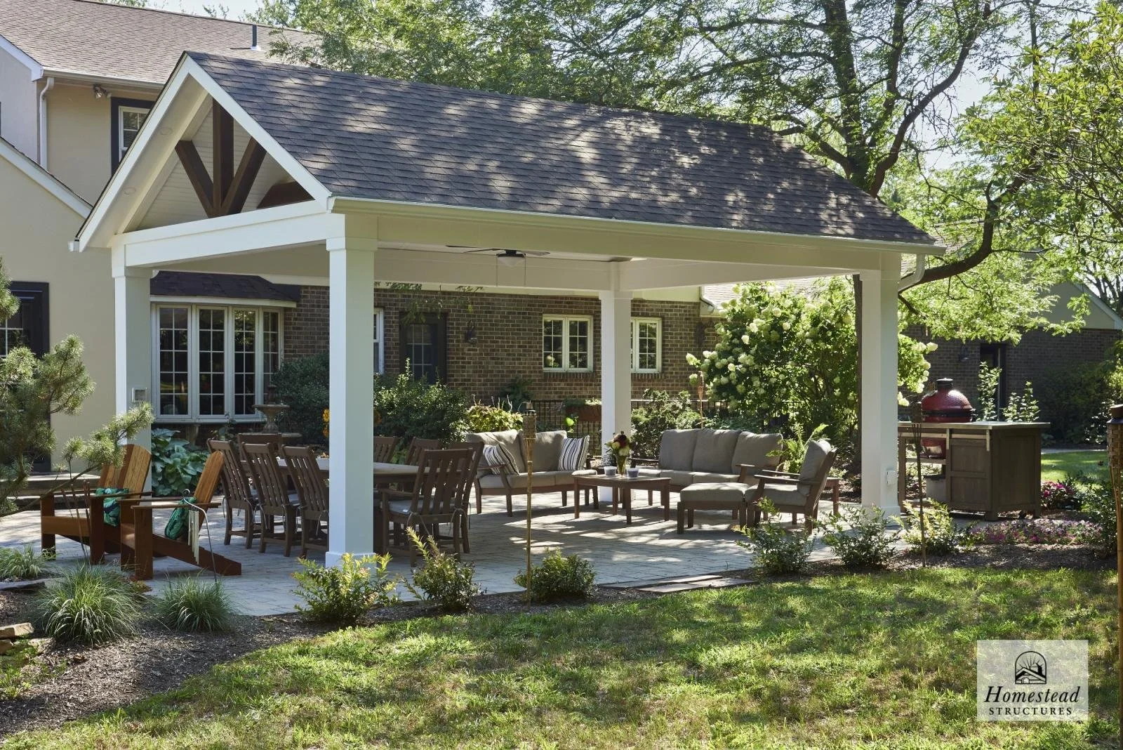 A backyard patio with outdoor furniture under a white pavilion, surrounded by greenery and a house in the background.