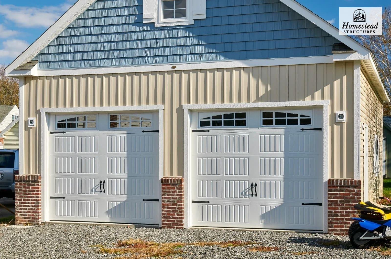 Double garage with two white doors, each featuring decorative window panels at the top, brick base supports, and black handles. Beige siding and blue roofing on the garage, with a small scooter visible on the right side.