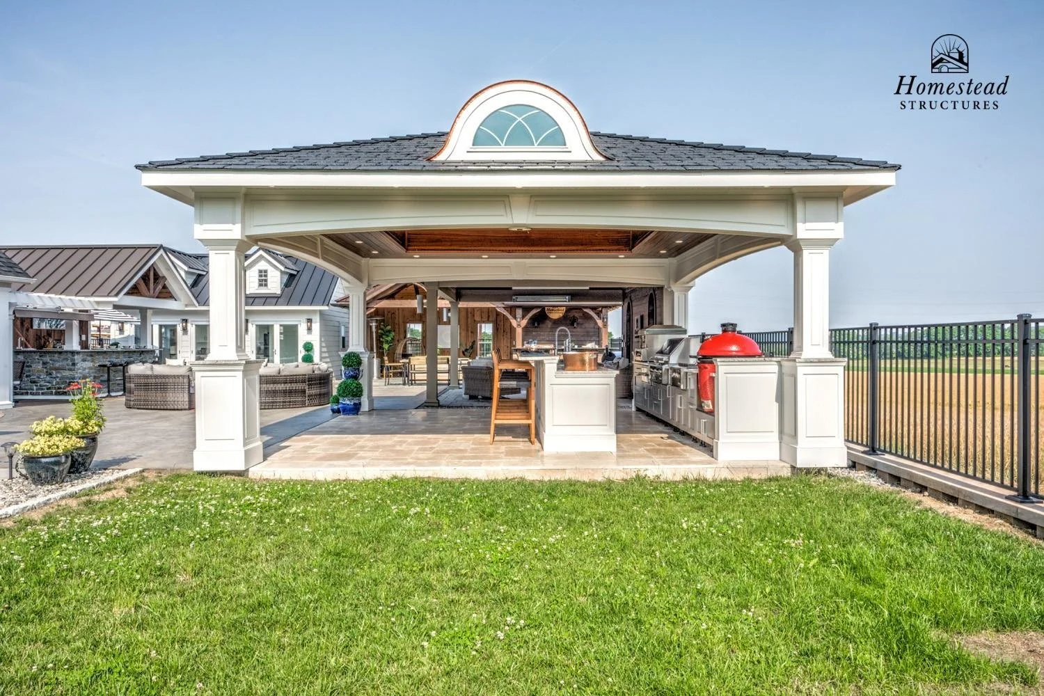 Outdoor kitchen with a covered patio, built-in grill, and seating area on a grassy backyard with neighboring houses in the background.