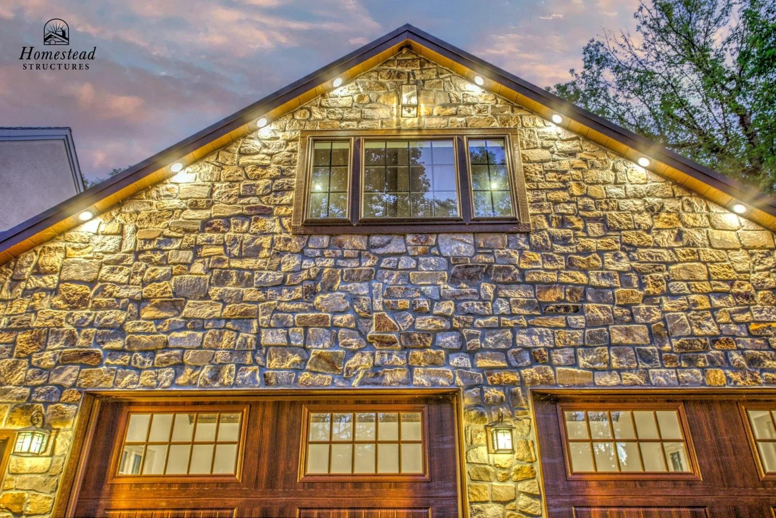 Exterior of a house with stone facade, illuminated at dusk, with wooden garage doors and a large window with multiple panes on the upper level, surrounded by trees.