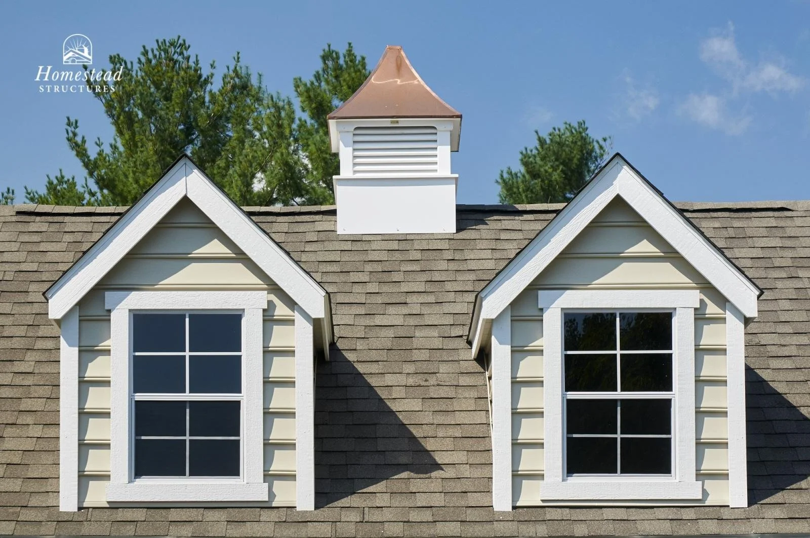 Close-up of a house roof with two dormer windows and a small white cupola vent against a blue sky with trees in the background.