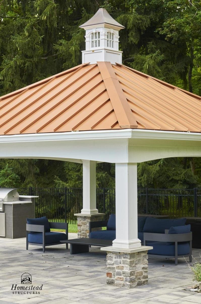 An outdoor pavilion with a copper-colored metal roof, white columns with stone bases, and corner seating with blue cushions, surrounded by greenery and a fenced yard.