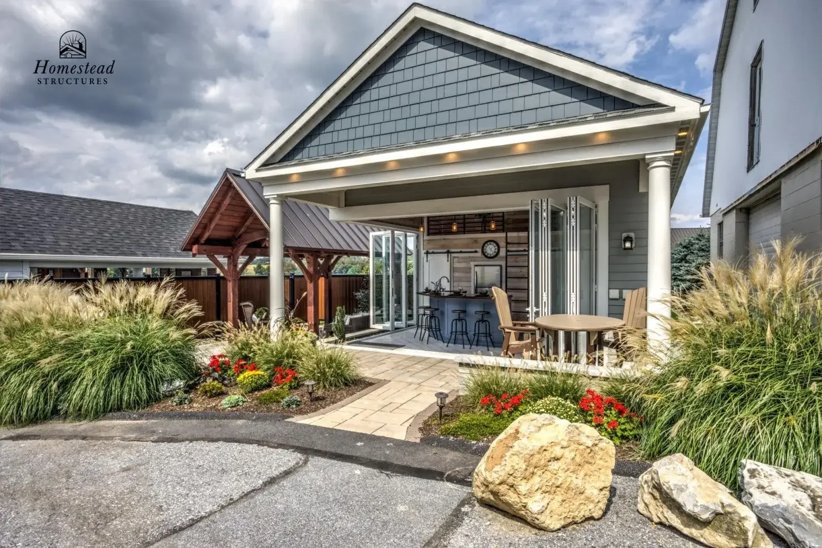Beautiful backyard patio with paver walkway, garden beds with ornamental grasses and flowers, a covered outdoor living area with a bar and stools, and an outdoor dining table with chairs. The house has gray siding, and there's a small wooden gazebo i