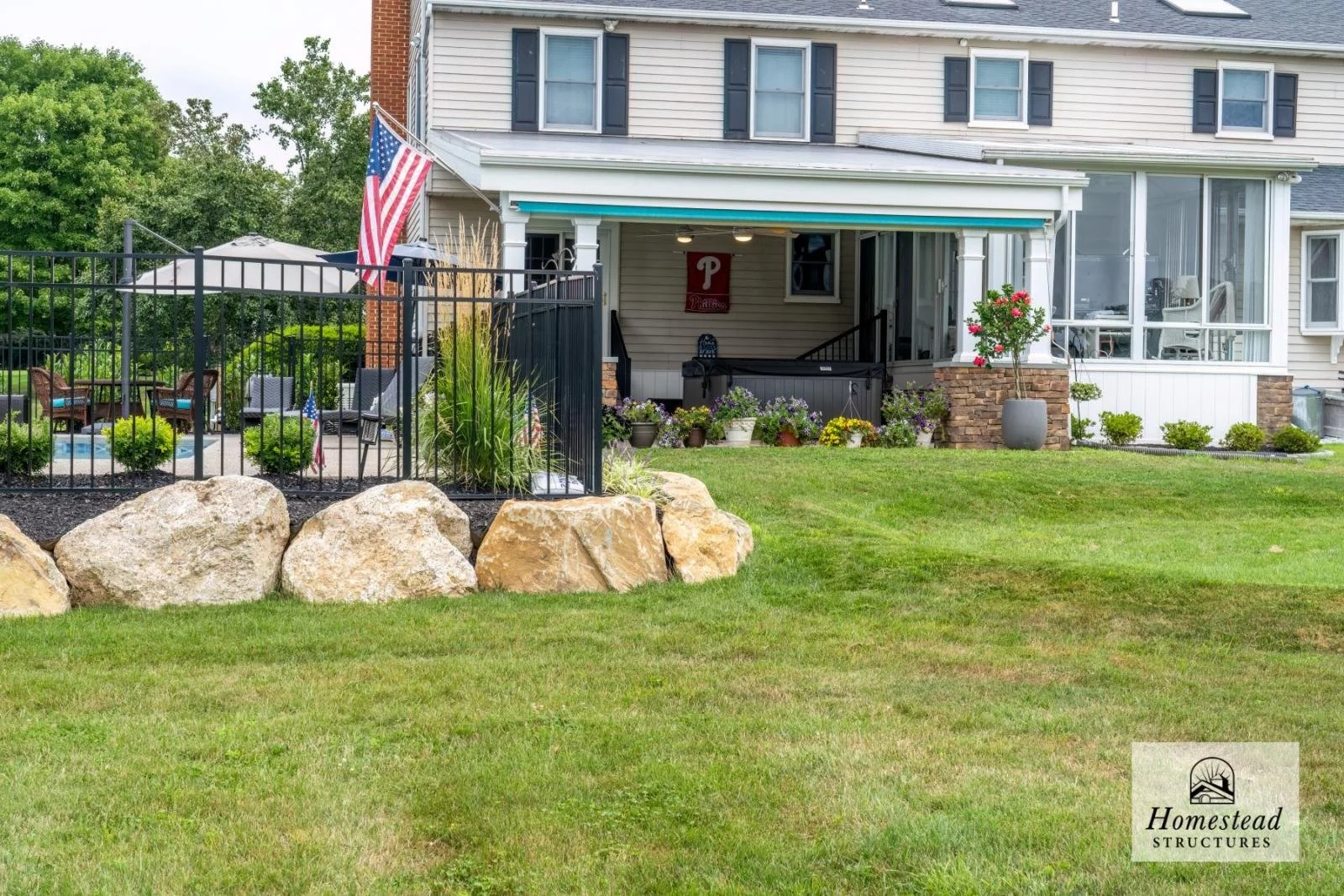 Backyard of a house with a patio area and a lawn. The patio is fenced with black metal and has outdoor furniture, umbrellas, and American flags. There are large rocks along the edge of the lawn, and various plants and potted flowers decorate the pati
