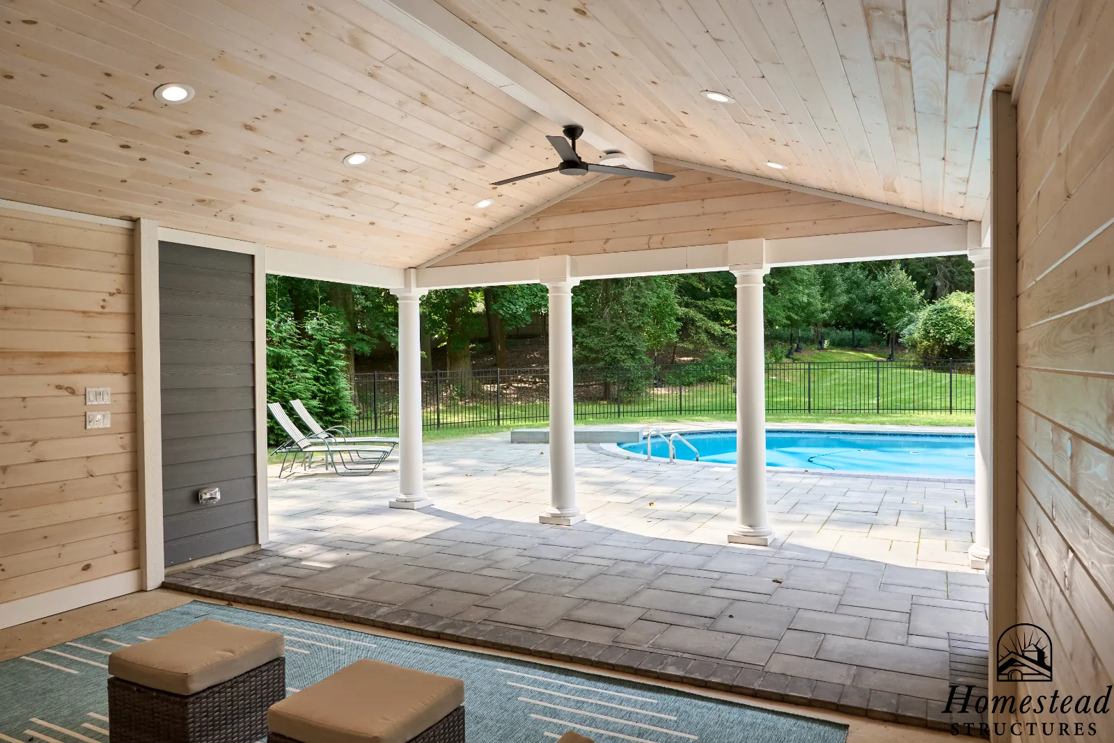 View from a covered porch looking out at a backyard with a pool, lawn, and trees, with patio chairs near the pool.