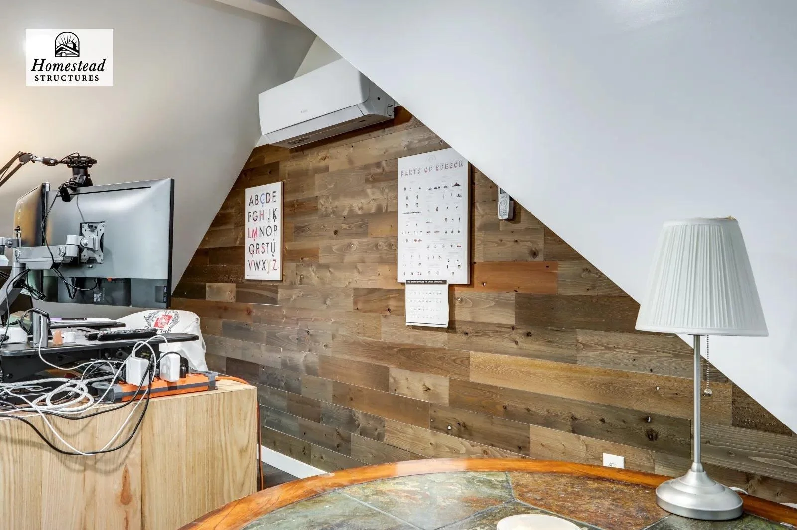 An office corner with a TV monitor, a wooden desk with wires, a white lamp on a wooden table, and educational posters on a wood-paneled wall.