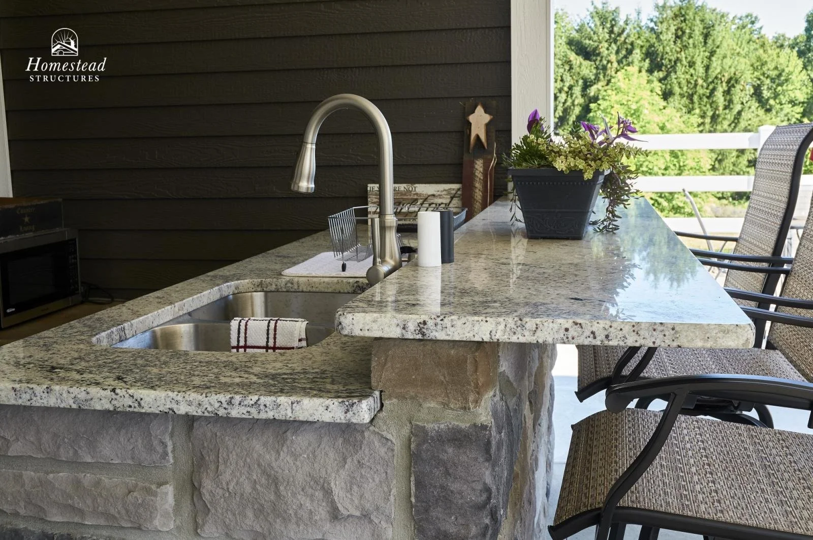 Outdoor kitchen with a granite countertop island featuring a sink, a stool with a woven seat, potted purple and white flowers, and decorative items, with a green wooded background.