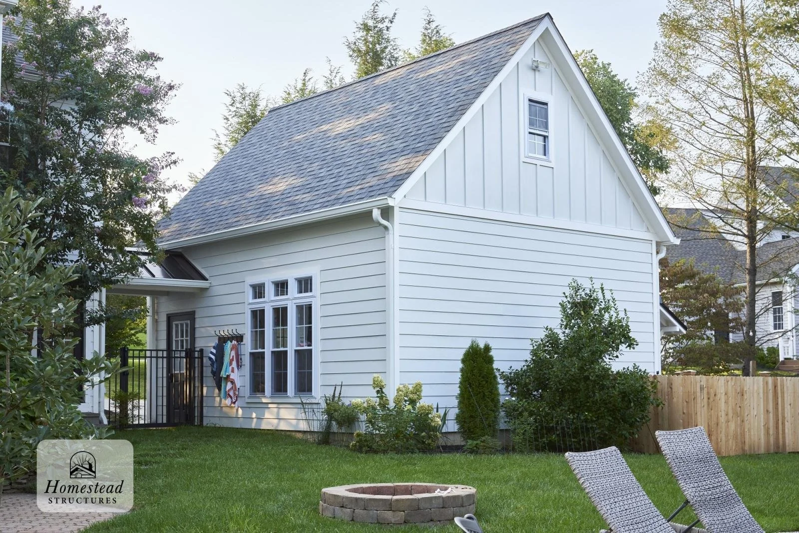 White house with gray shingle roof in a backyard with green grass, bushes, and trees, fenced with a wooden fence and black gate, lounge chairs and a fire pit.