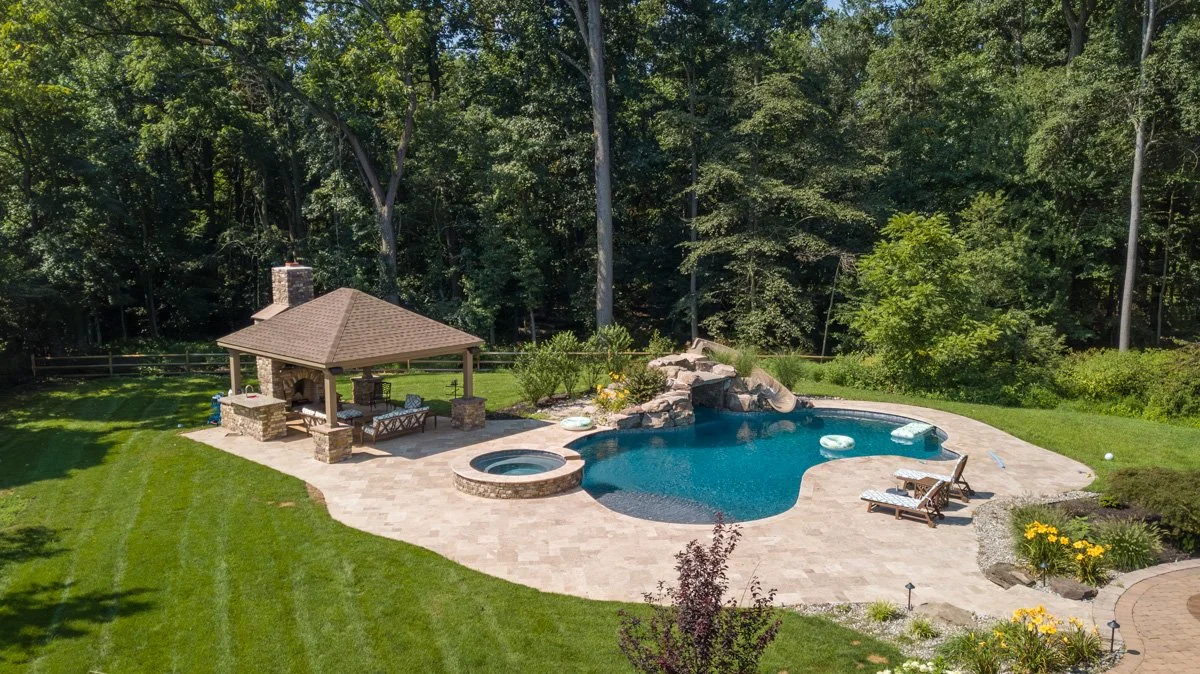 Rear view of a backyard featuring a swimming pool with rock waterfall, a hot tub, and a covered outdoor pavilion with seating, surrounded by green lawn and trees.