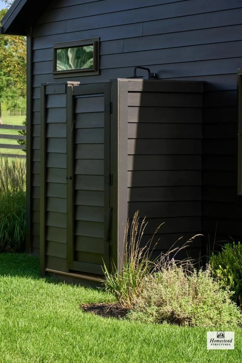 A small black shed with a double door, situated next to a blue house with horizontal siding. There is a small rectangular window above the shed and grass with some plants in front of it.