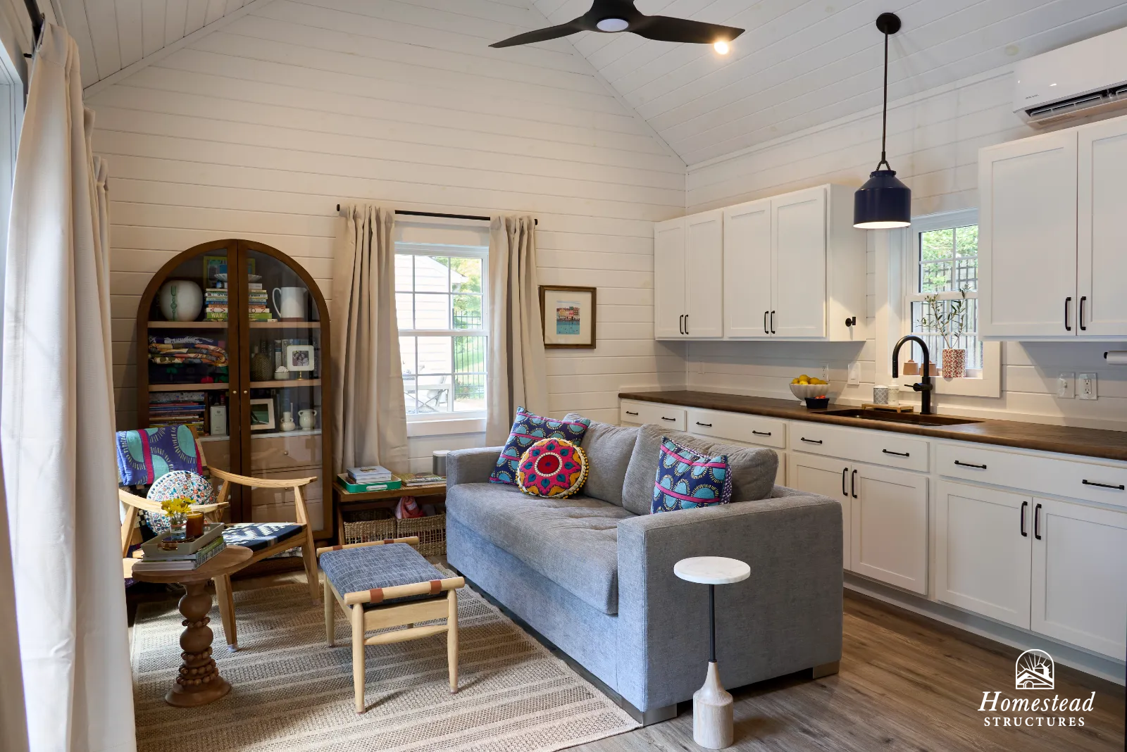 Living room with white shiplap walls, a gray sofa with colorful patterned pillows, a wooden side table, a wooden armchair with a blue cushion, a round wooden coffee table with books and a flower vase, a woven rug, a bookshelf, and white kitchen cabin