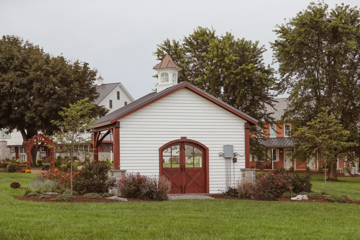 White barn with brown trim and double doors, surrounded by green grass, trees, and landscaped flower beds, with a neighboring house and trees in the background.