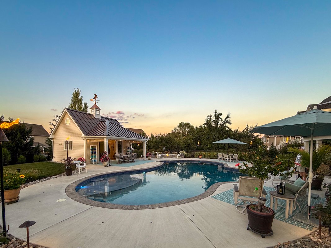 A backyard with a kidney-shaped swimming pool, surrounded by patio furniture, umbrellas, and potted plants, at sunset.
