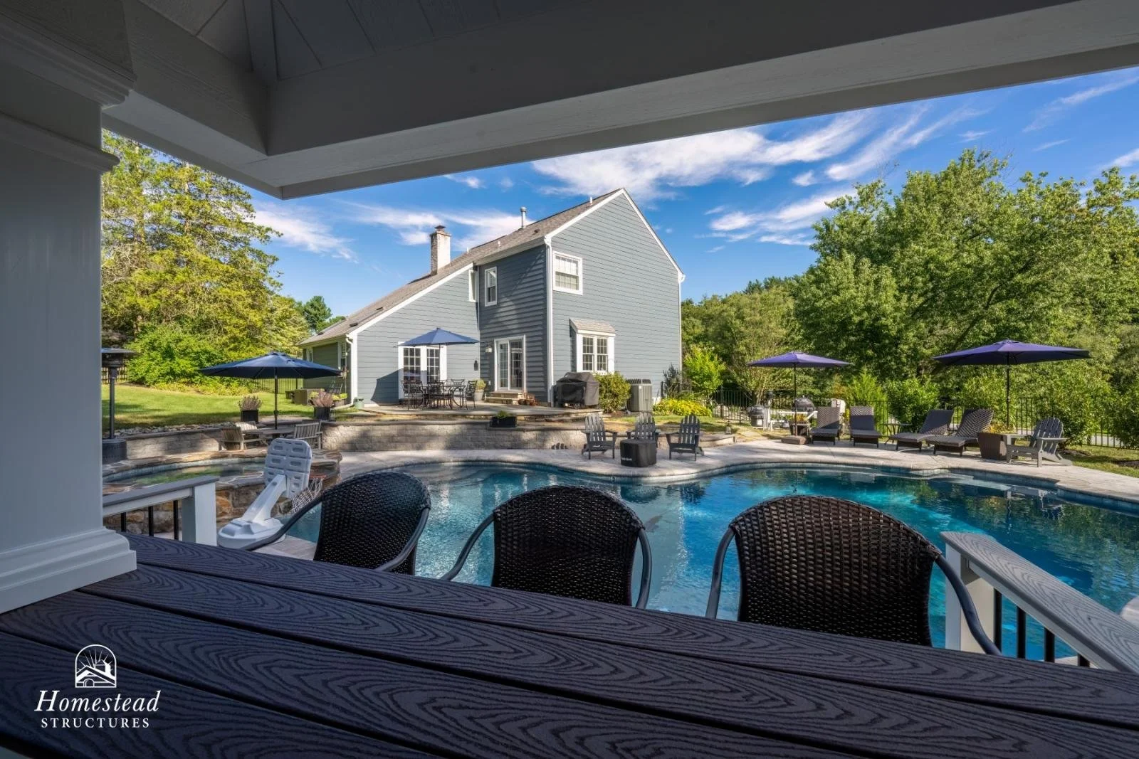 View of a backyard with a swimming pool, covered patio, outdoor seating and lounge chairs, surrounded by trees, with a blue house and a partly cloudy sky in the background.
