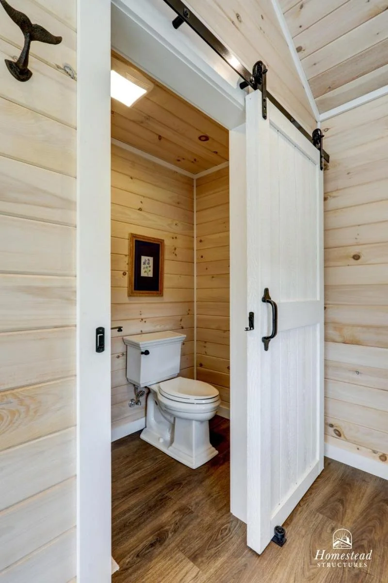 A small bathroom with wooden paneled walls, a white toilet, a framed picture on the wall, and a sliding barn door.