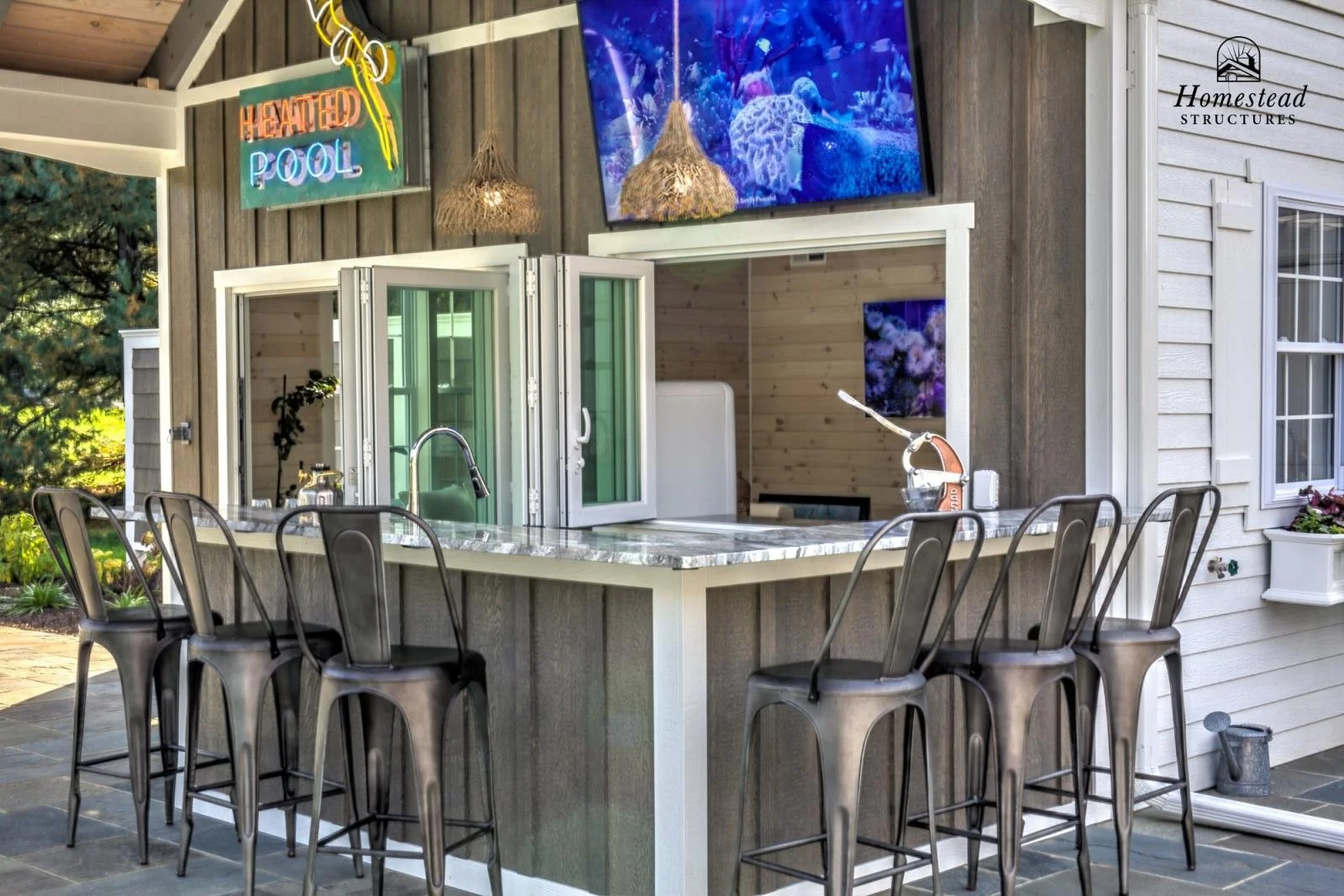 Outdoor kitchen with a marble counter, bar stools, and a window revealing an indoor space. A large flat-screen TV is mounted above, displaying an underwater scene. There are two hanging pendant lights and a neon sign reading "Heated Pool."