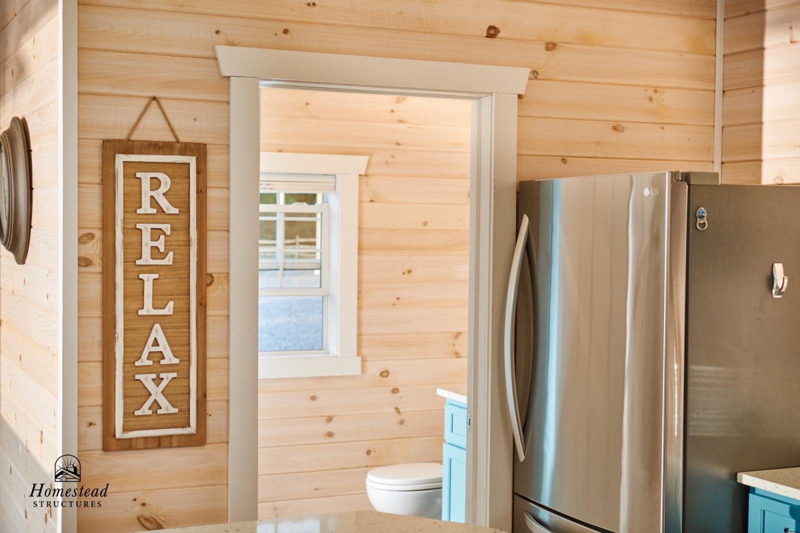 Interior view of a rustic home with light wood paneling on walls, a kitchen counter in the foreground, a stainless steel refrigerator on the right, and a doorway leading to a bathroom with a toilet and a window in the background. A decorative sign sa