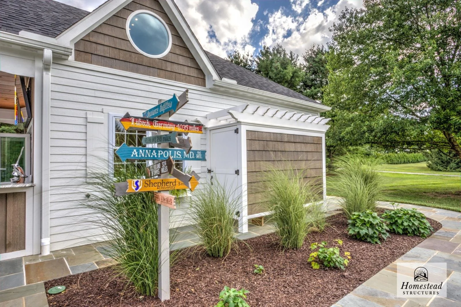 Colorful garden signpost with directional arrows pointing to multiple locations in a landscaped yard next to a house with white siding and brown shingles, under a cloudy sky with trees in the background.