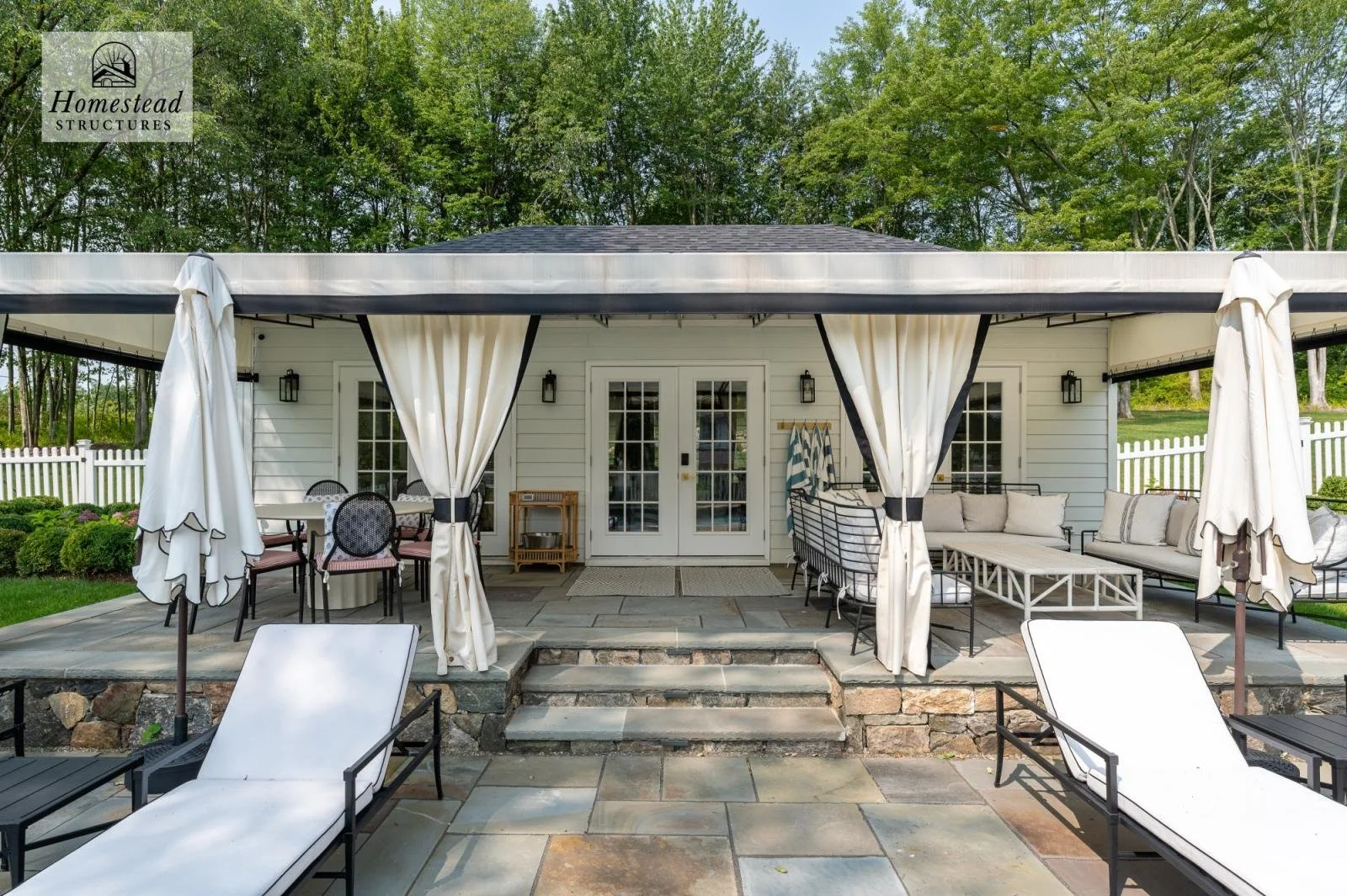 Outdoor patio with white cushioned lounge chairs, umbrellas, and seating area in front of a white house with double glass doors, surrounded by green trees and a white picket fence.