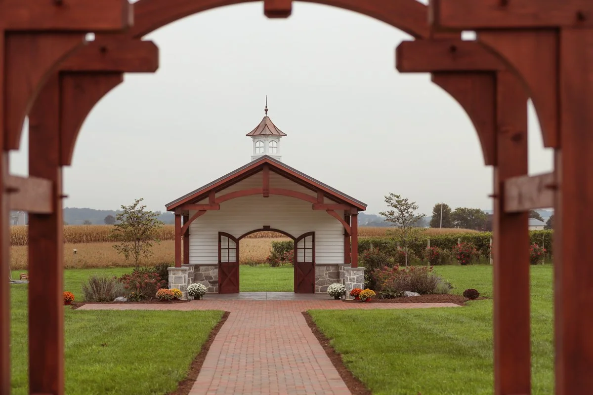 A small white building with red trim, a steeple on top, and double doors, viewed through a wooden archway with a brick pathway leading to the entrance, surrounded by a well-manicured lawn and garden on an overcast day.