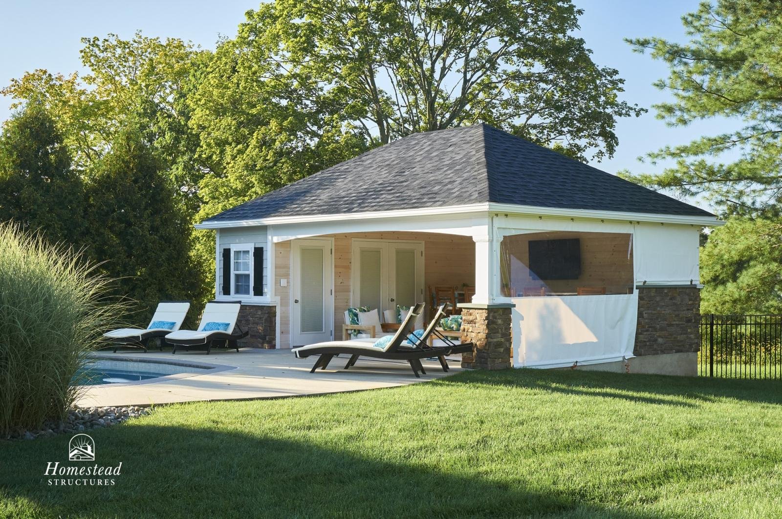 A small backyard with a house featuring a gray shingle roof, brick and white siding, and a screened porch with seating. There are lounge chairs by a swimming pool, and the yard is lush with green grass and trees in the background.