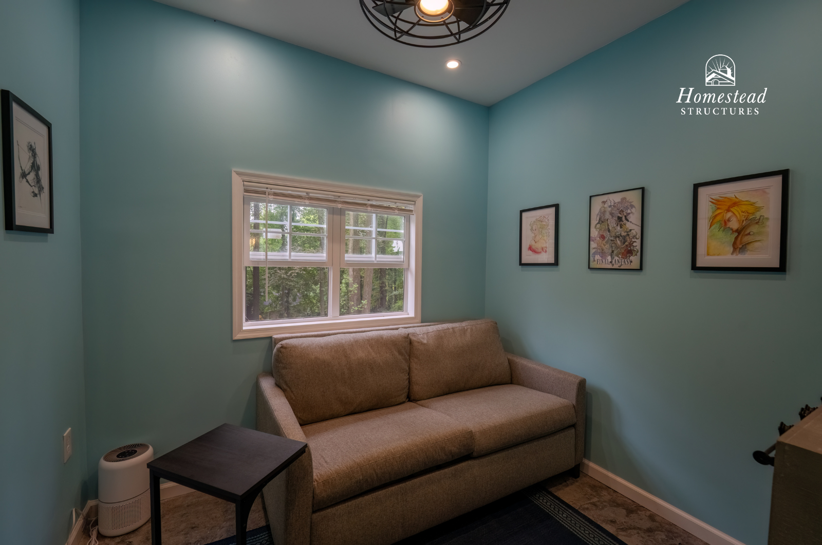 Small living room with light blue walls, a beige sofa, a small black side table, framed artwork on the walls, a window with blinds, ceiling light fixture, and wood flooring.