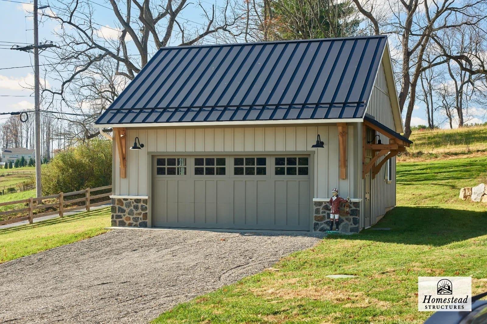 A beige and gray detached garage with a metal roof, stone accents at the base, and overhead lighting, situated on a gravel driveway with a lawn and trees in the background.