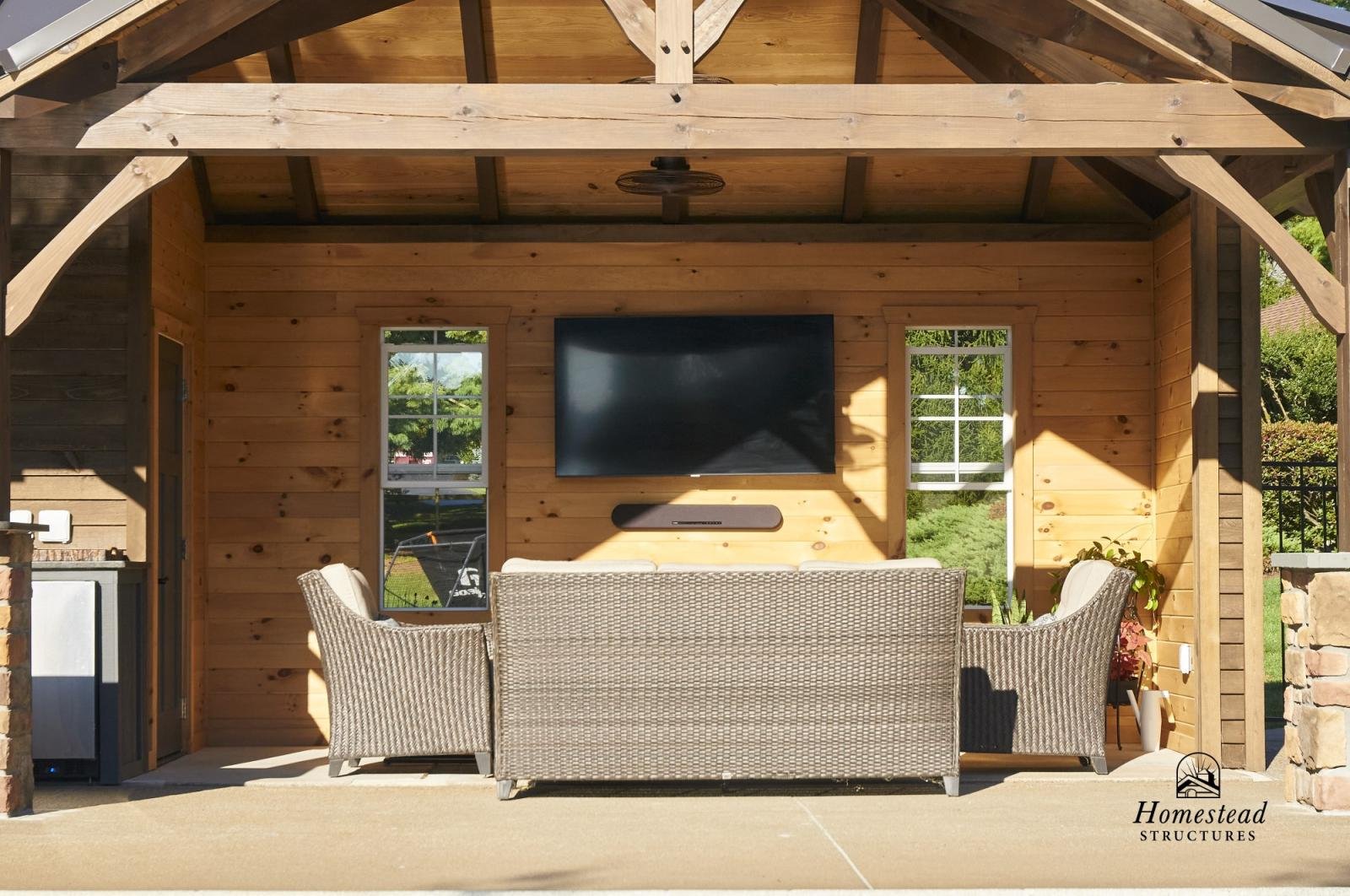 An outdoor patio area with a wooden wall and roof structure. There is a large flat-screen TV mounted on the wall, a soundbar below it, and a wicker sofa with white cushions facing the TV. To the right, there is a potted plant. The area is shaded, wit
